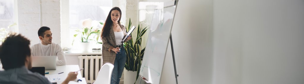Eine junge Frau steht vor einem Whiteboard und präsentiert Informationen. Zwei Männer sitzen an einem Tisch und hören ihr aufmerksam zu. | Une jeune femme se tient devant un tableau blanc et présente des informations. Deux hommes sont assis à une table et l'écoutent attentivement. | A young woman stands in front of a whiteboard and presents information. Two men are sitting at a table and are listening to her attentively.